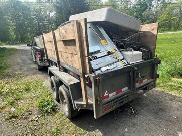 Loaded dump trailer filled with scrap wood and metal on a gravel road beside trees.