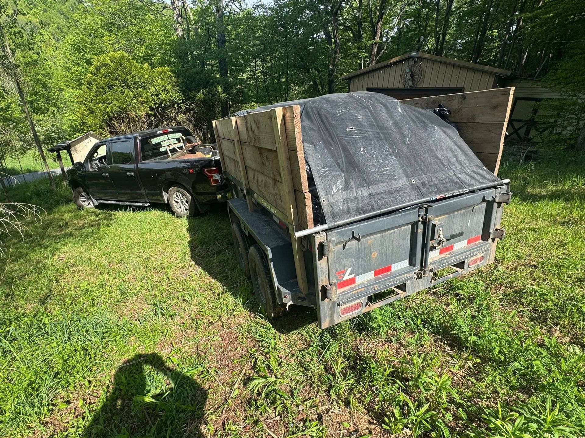 Small utility trailer loaded with wood in a grassy yard beside a black pickup truck and shed.
