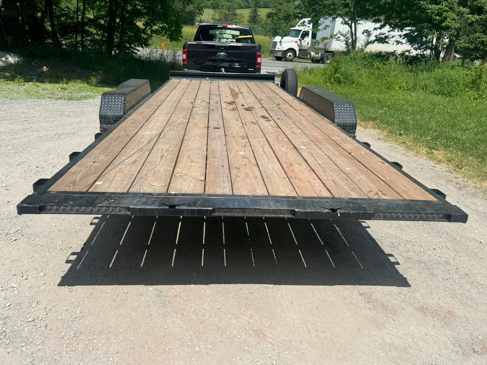 Flatbed trailer with wooden deck parked on a road, with a pickup truck ahead in the background.