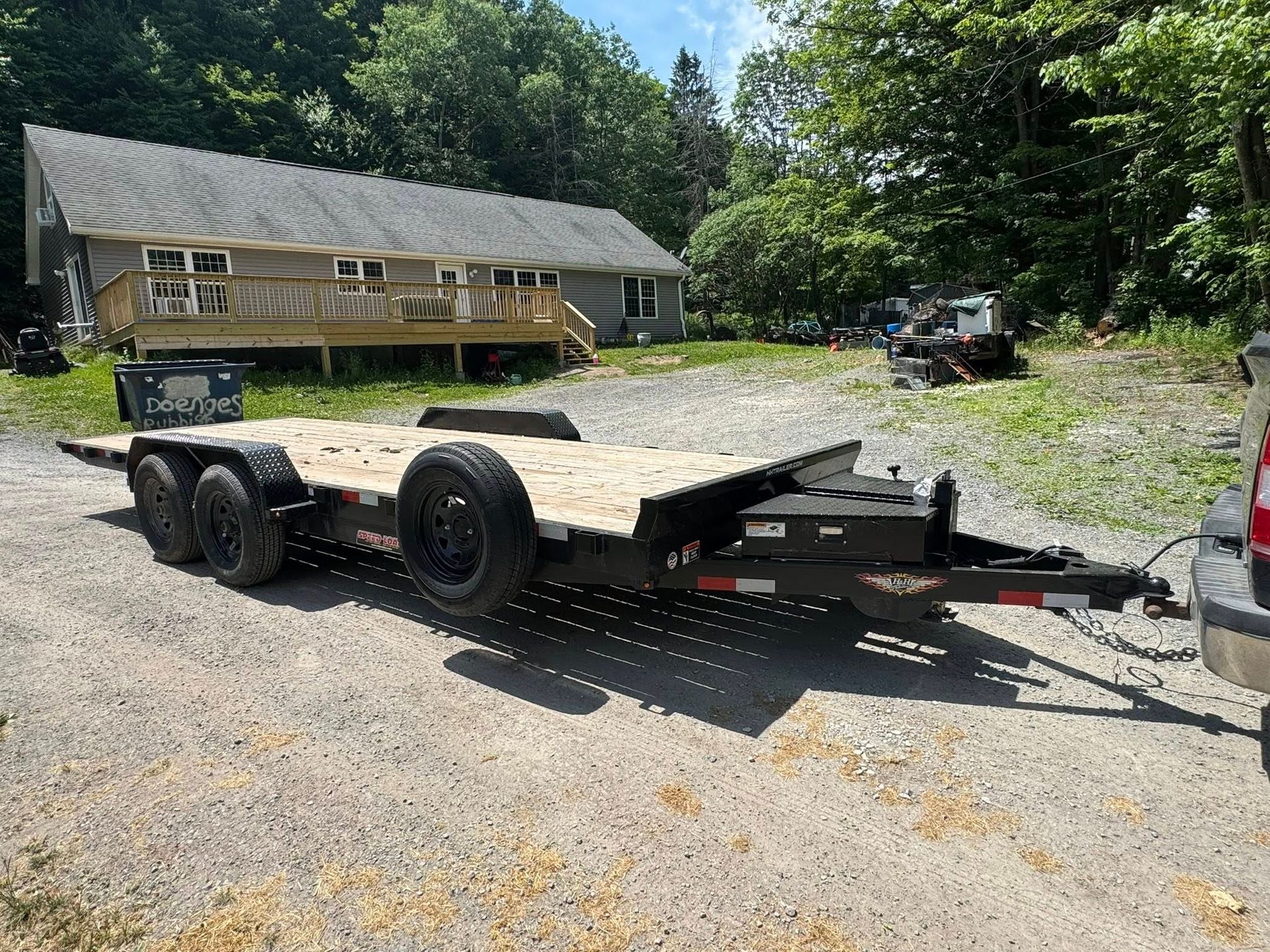 Flatbed trailer parked on a gravel driveway beside a small cabin and trees.