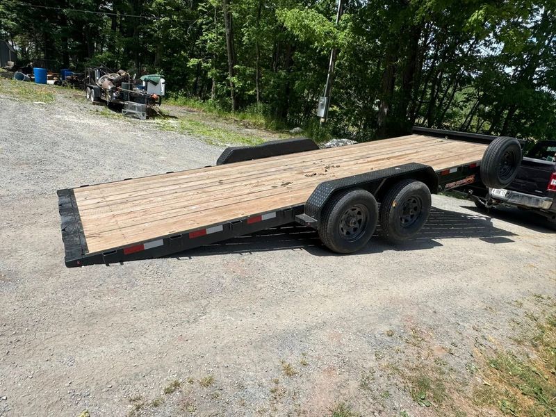 Black flatbed car trailer parked on a gravel lot near trees, with wooden deck and dual axles.