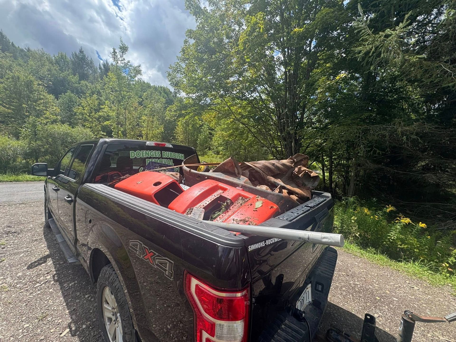 Black pickup truck bed loaded with orange ATV parts, debris, and a metal bar, parked near trees.
