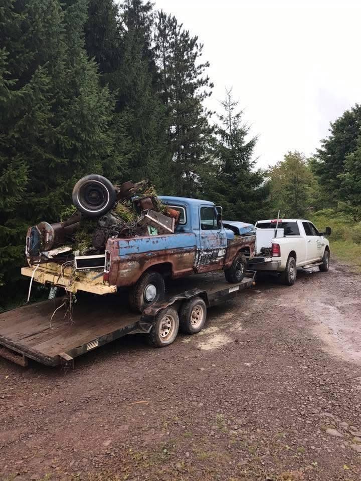 White pickup truck towing a trailer with a rusty blue truck loaded with scrap metal in a forest.