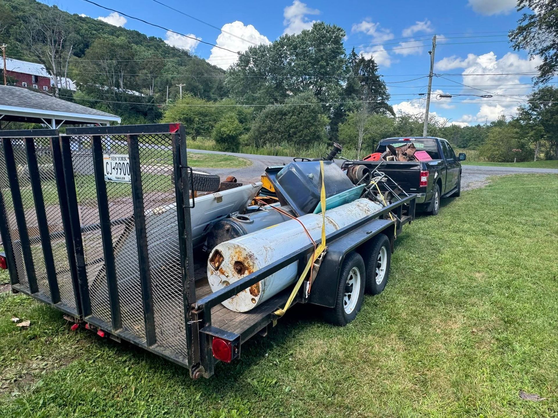 Black truck towing a trailer loaded with metal scraps, parked on grass near a road, cloudy sky.