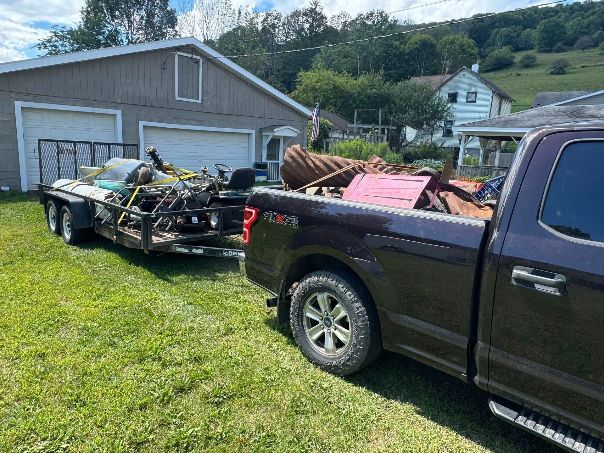 A burgundy pickup truck towing a trailer loaded with junk, parked on a grassy lawn near a garage.