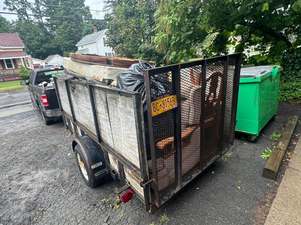 Trailer filled with debris parked next to a green dumpster. Black truck in background, houses and trees visible.