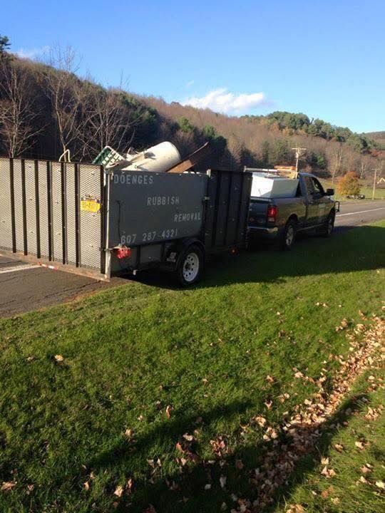 Truck towing a trailer filled with debris near a chain link fence and green grass. Blue sky and trees in background.