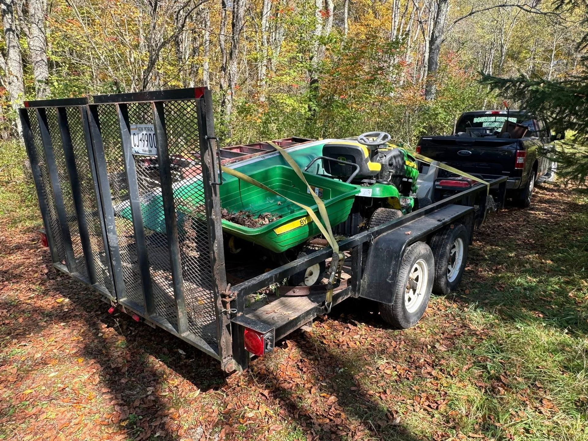 Trailer with a green riding lawnmower and debris, hitched to a black pickup truck, in a wooded area.