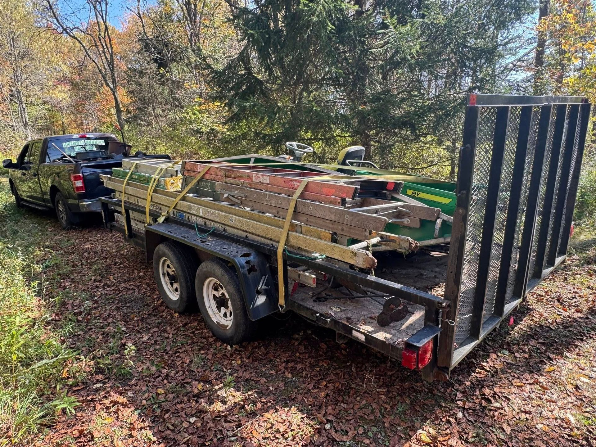 Truck towing a trailer loaded with ladders and a green lawn mower on a dirt road.