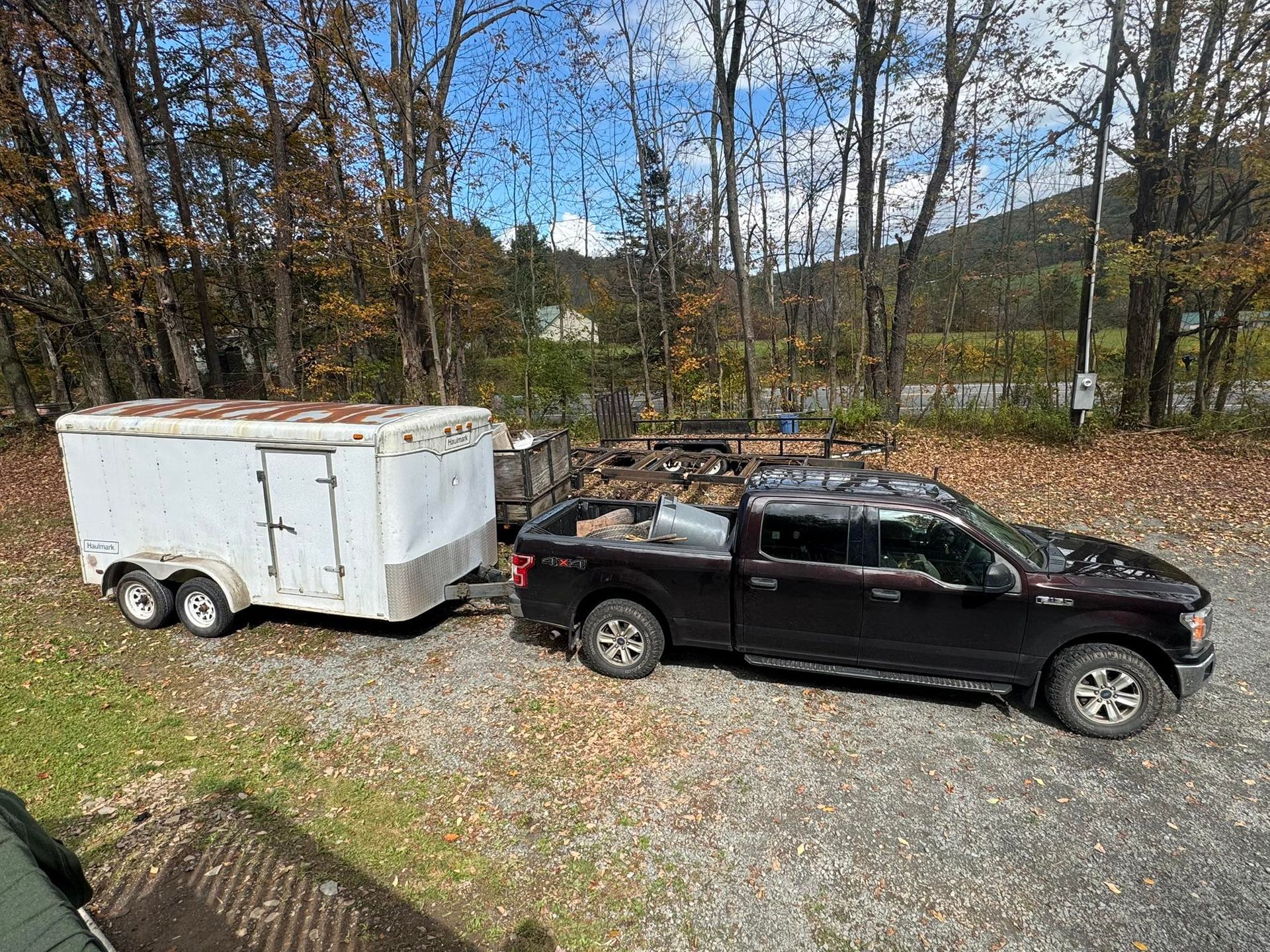 Dark pickup truck towing a white enclosed trailer on a gravel driveway, trees in the background.