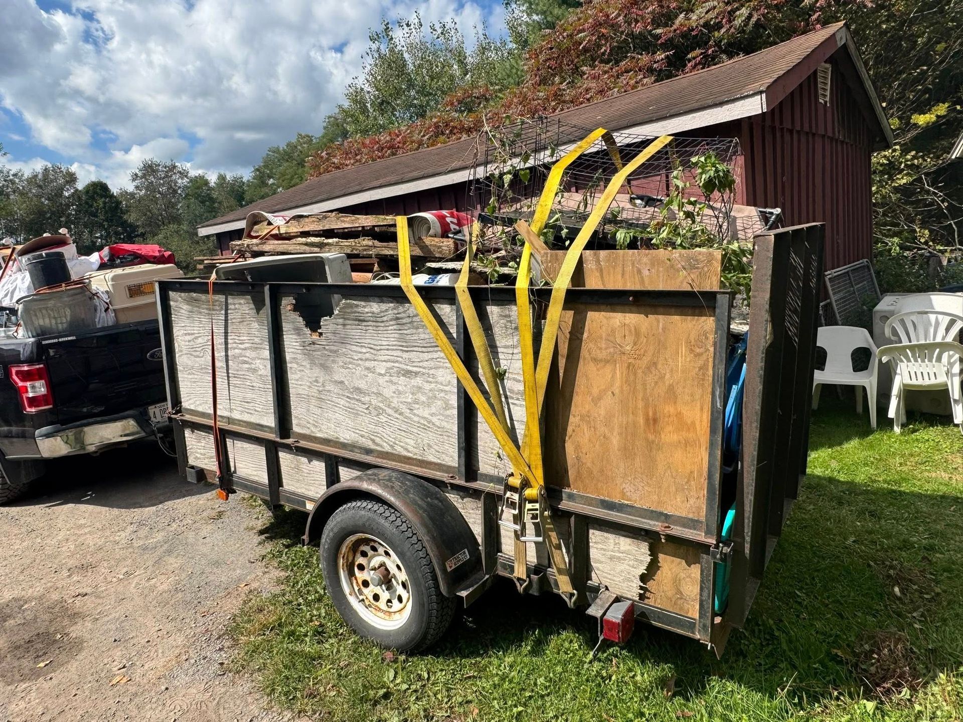 Trailer loaded with wood and debris, secured with yellow straps, parked on gravel.