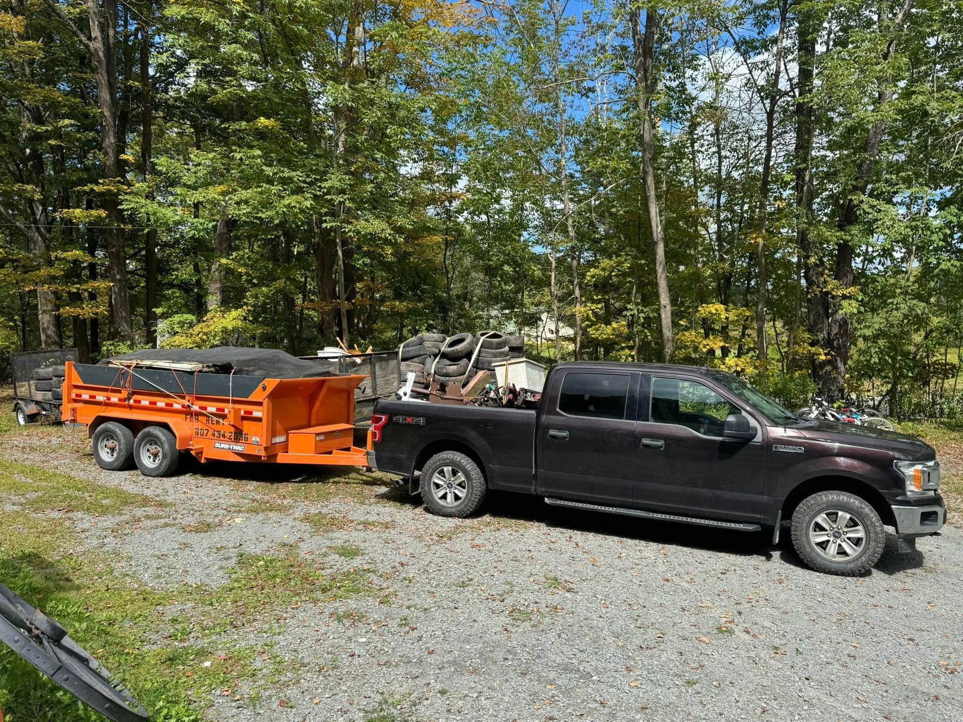 Dark truck towing orange trailer on gravel, trees in background.