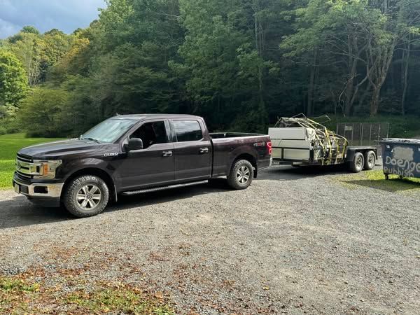 Dark pickup truck towing a trailer on a gravel driveway, wooded background.