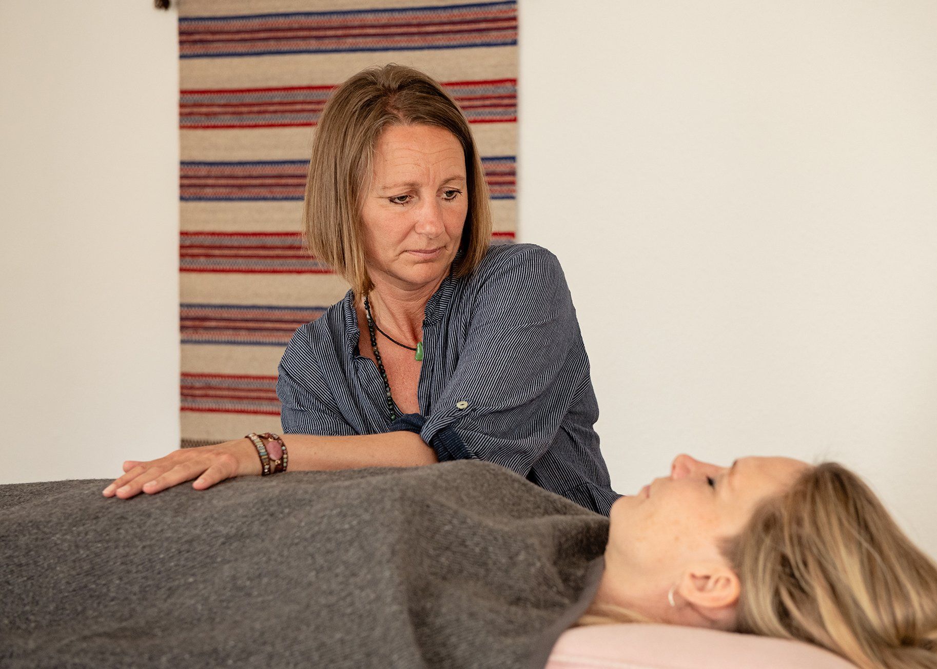 Kvinde står ved siden af ​​en tildækket person på en massagebriks i et roligt rum med vævet vægdekoration.