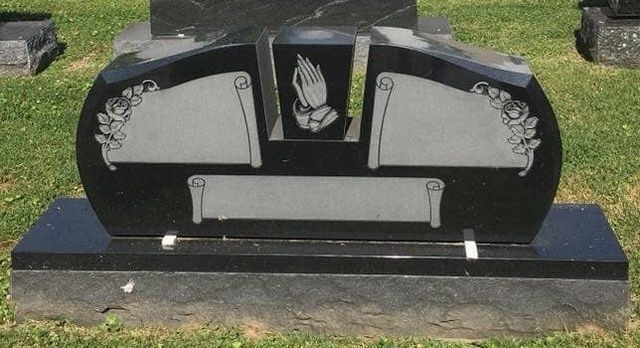 A black gravestone with praying hands on it is in a cemetery.