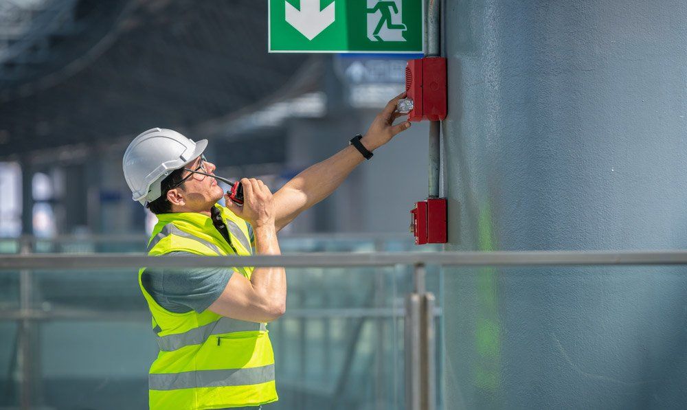 Construction Worker in Hard Hat and Vest Activates a Fire Alarm, Near an Exit Sign — Hunter Fire & Safety in Edgeworth, NSW