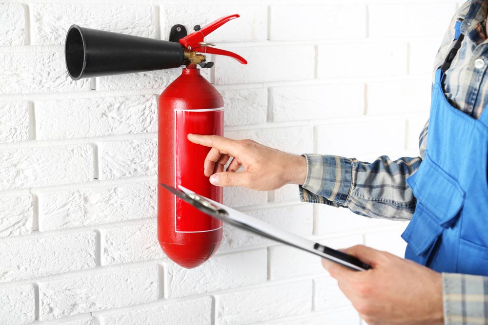 Person Inspecting a Red Fire Extinguisher on a White Brick Wall — Hunter Fire & Safety in Central Coast, NSW
