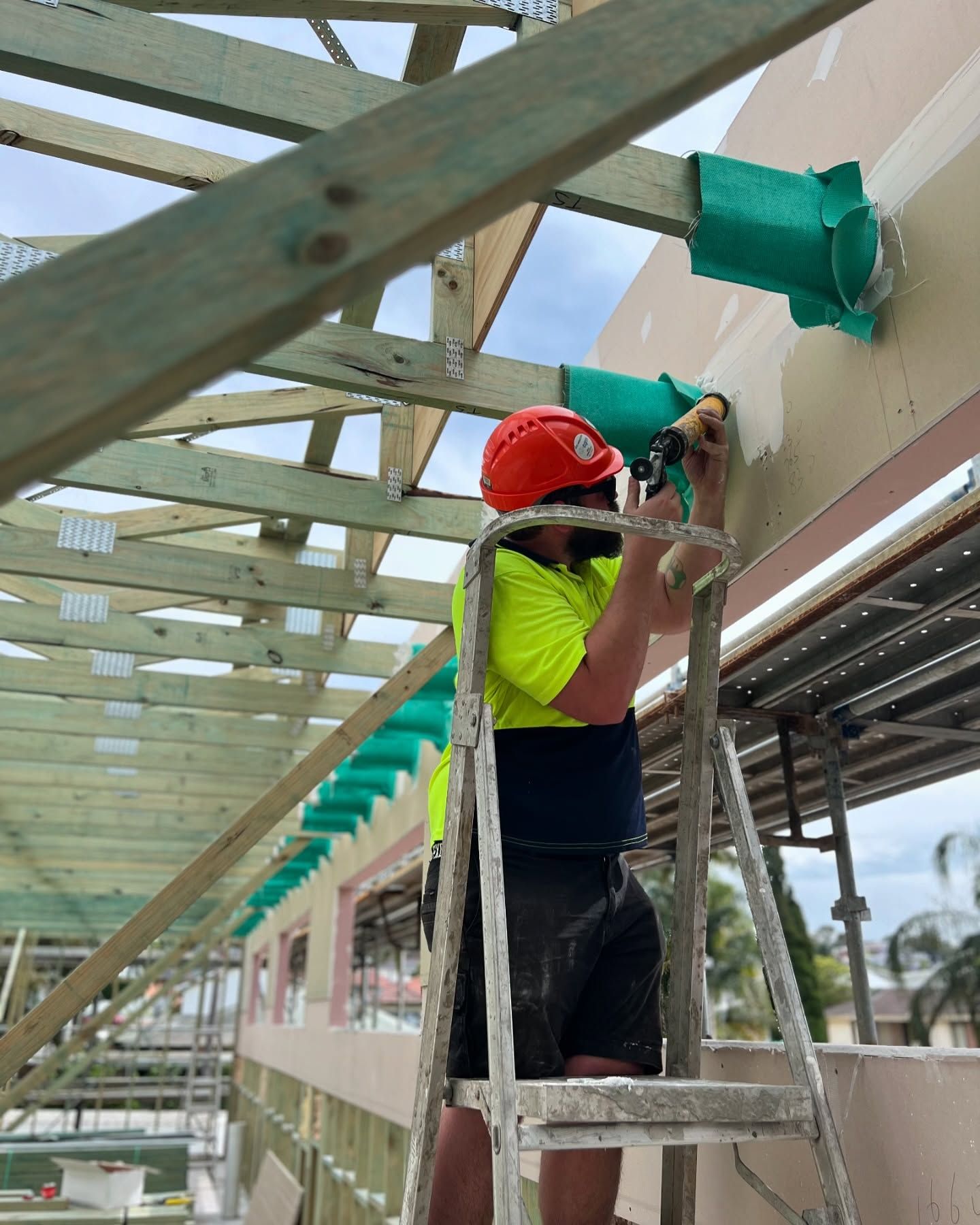 Construction Worker in Safety Gear on Ladder, Using a Tool on a Wall — Hunter Fire & Safety in Edgeworth, NSW