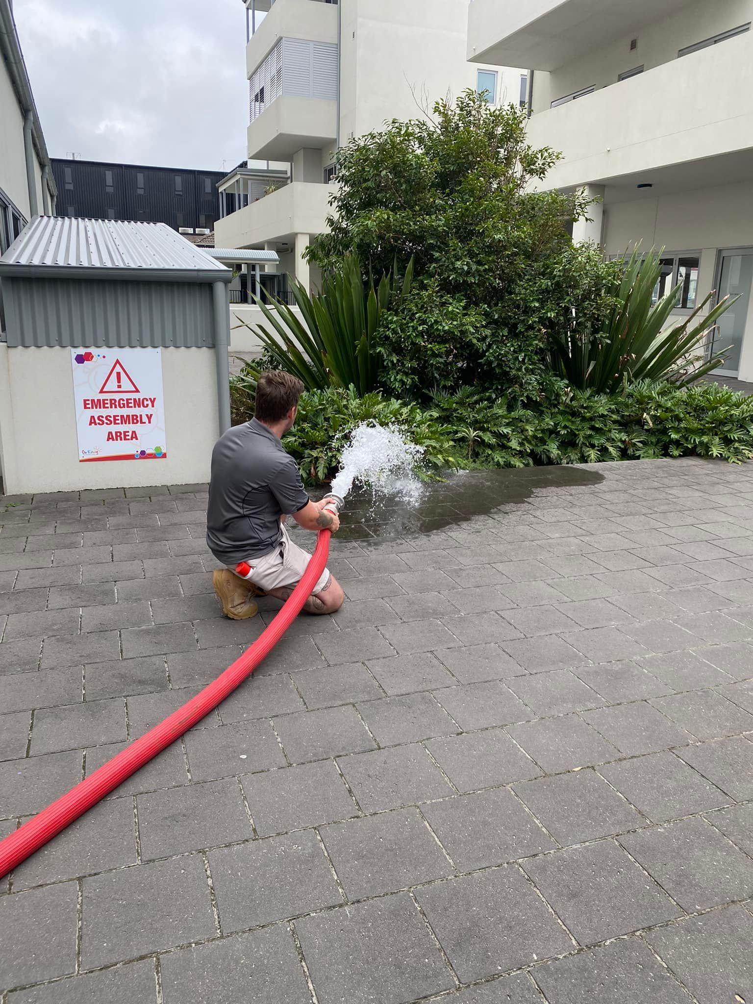 Man Kneeling, Spraying Water From a Red Hose Onto a Paved Area — Hunter Fire & Safety in Edgeworth, NSW