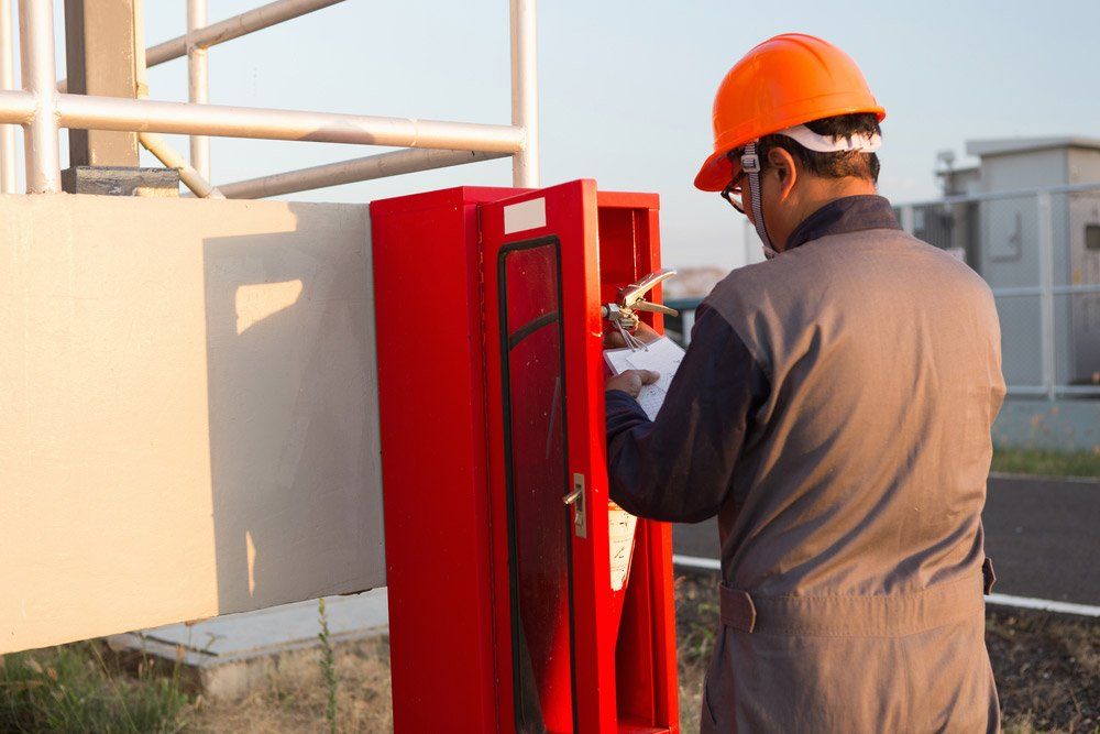 Man in Orange Hard Hat Inspecting a Red Fire Extinguisher Cabinet Outdoors — Hunter Fire & Safety in Central Coast, NSW