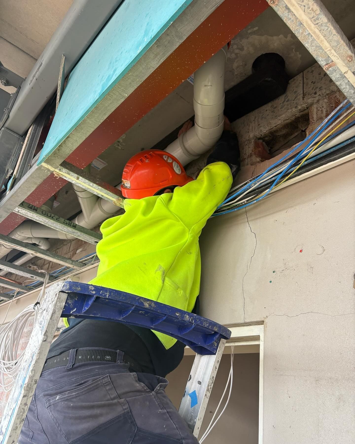 Construction Worker in a Yellow Jacket and Orange Helmet, Working on Pipes Above — Hunter Fire & Safety in Edgeworth, NSW