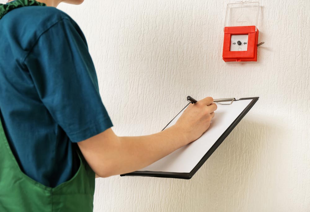 Person in Green Overalls Inspecting a Red Fire Alarm — Hunter Fire & Safety in Edgeworth, NSW
