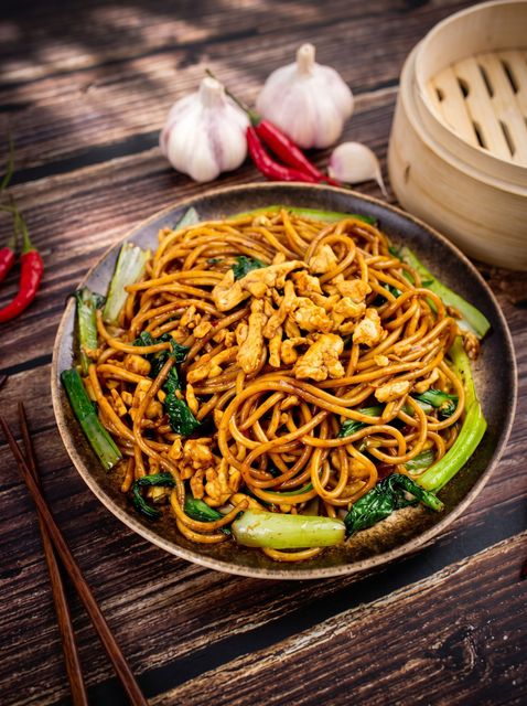Stir-fried noodles with vegetables in a pan, garnished with garlic and red chilies on a rustic wooden table