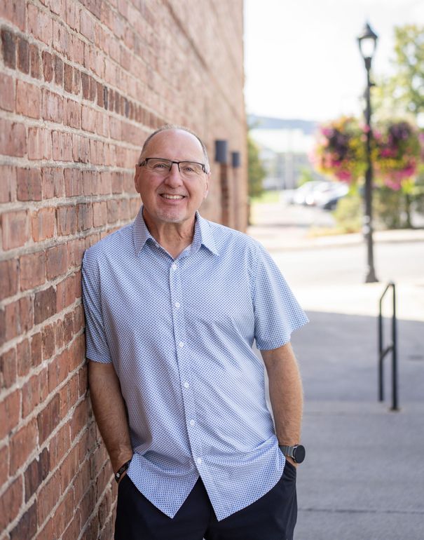 Man in patterned shirt leans against brick wall, smiling. Outdoors, street, sunny.