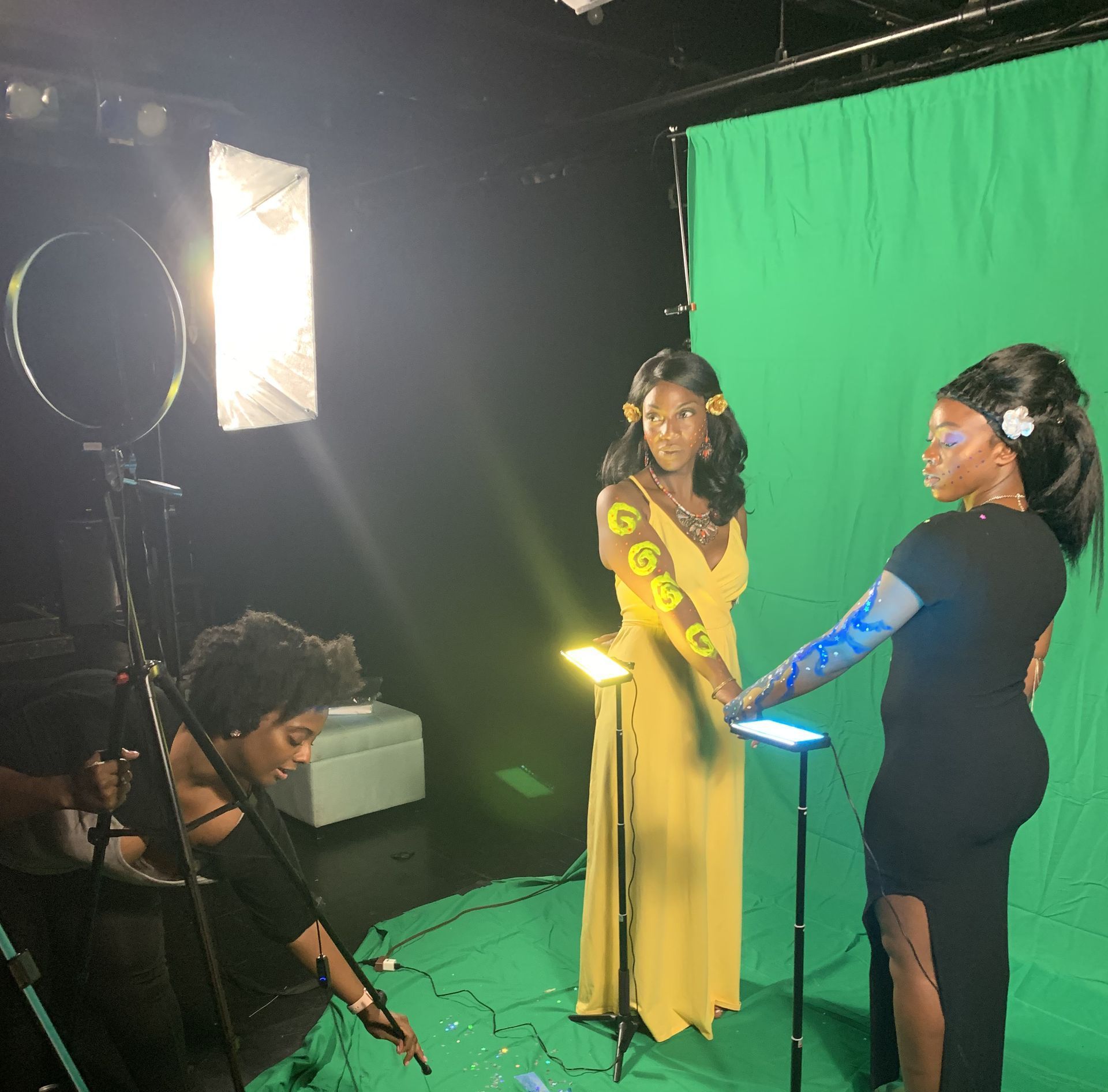 Film set with two people beside a green screen and studio lights, one holding a tablet while another adjusts equipment