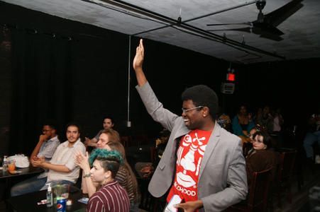 Man in gray blazer and red shirt raises his hand at a crowded indoor event