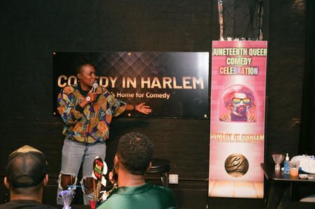 Comedian performing on stage at Comedy in Harlem, with a colorful event banner beside him.