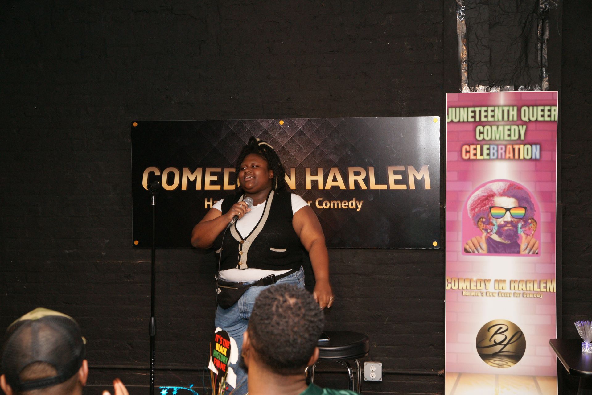 Comedian on stage at Harlem comedy club, speaking into a microphone beside a Juneteenth Queer Comedy Celebration banner