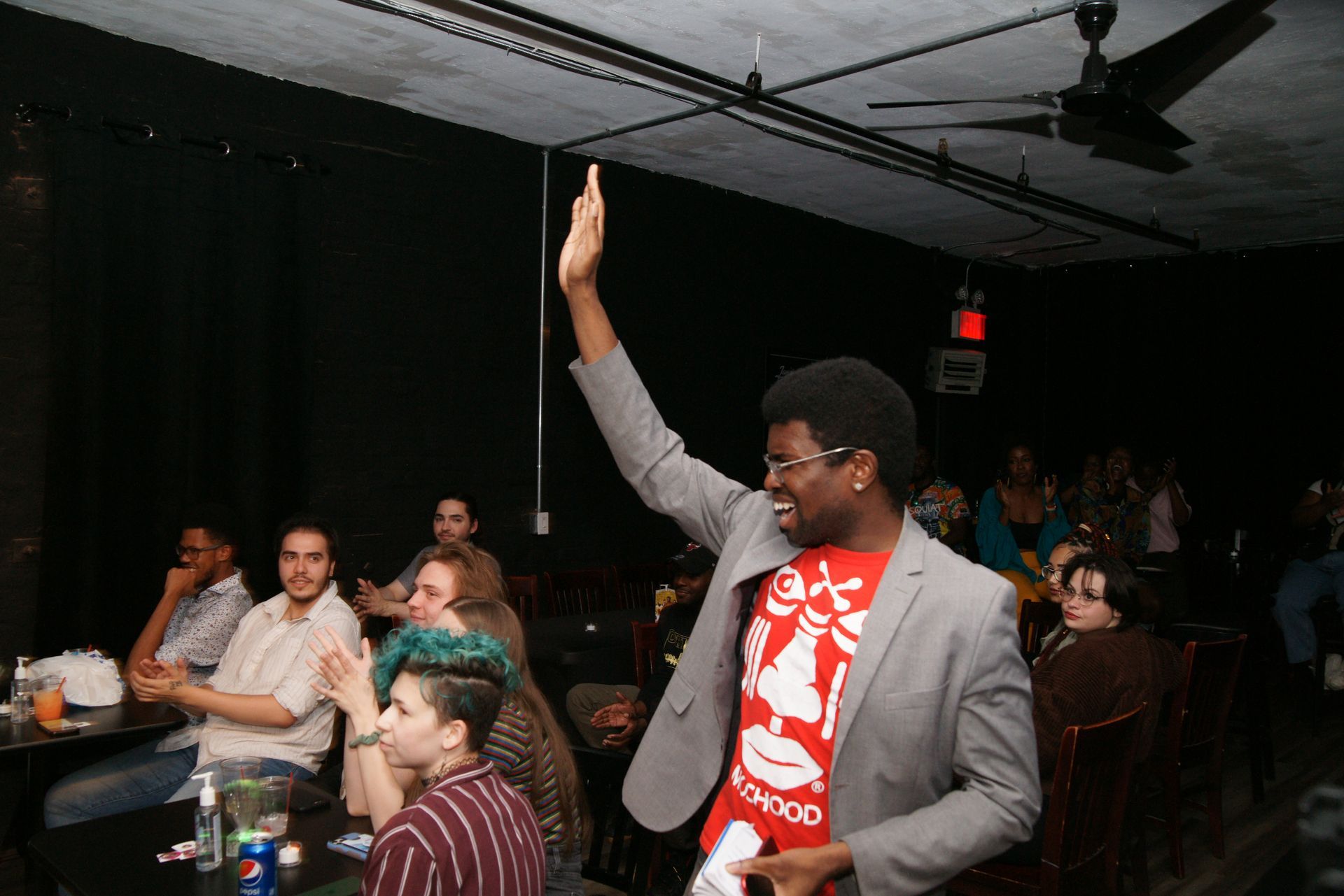Person in gray blazer raises a hand in a dim auditorium, wearing a red T-shirt with a white graphic.