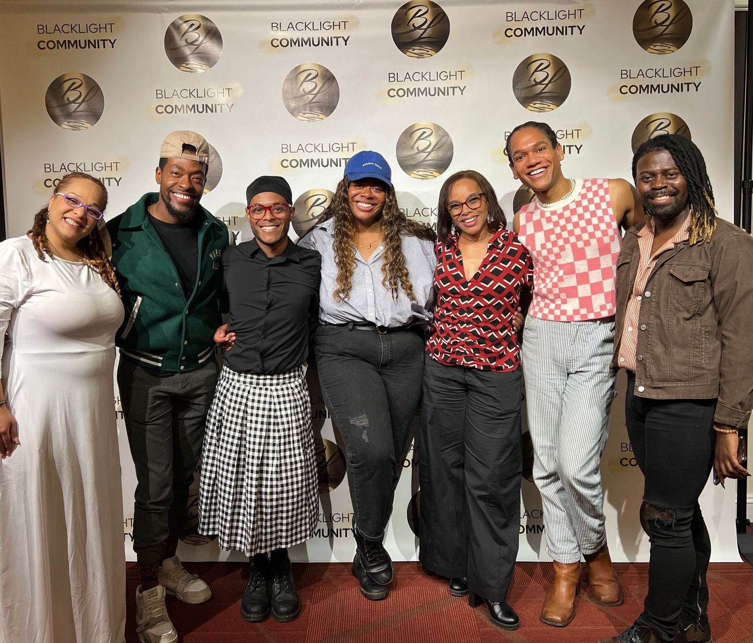 Seven people posing together indoors in front of a branded backdrop, smiling and dressed in colorful outfits.