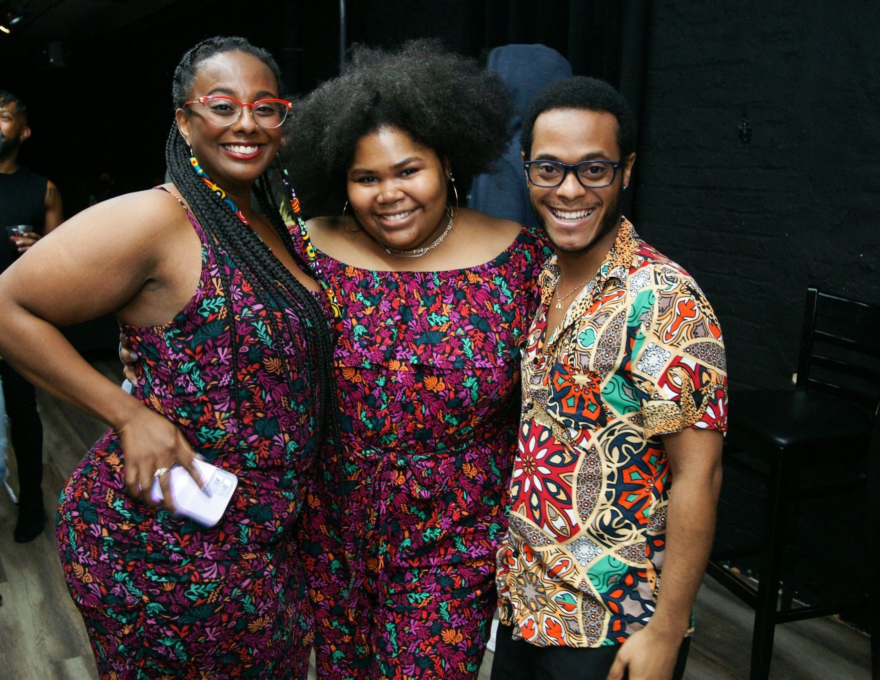 Three people smiling and posing together indoors, wearing colorful patterned clothes.