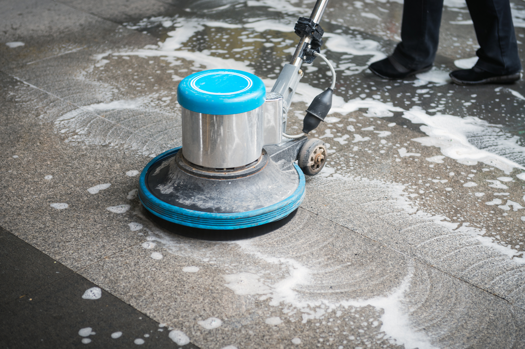 Person cleaning a concrete surface with a blue and silver floor scrubber, creating suds.