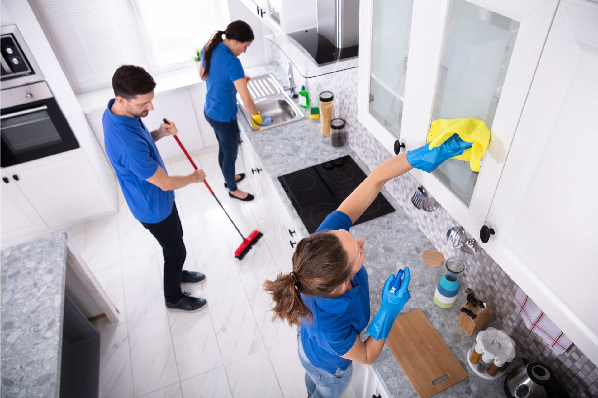 Three people cleaning a white kitchen: sweeping, wiping counters, and cabinet doors.