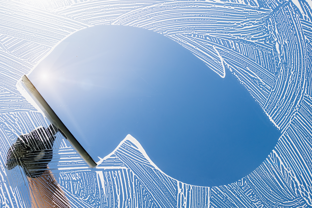 Hand cleaning a window with a squeegee, leaving a clear patch against a soapy, blue sky.
