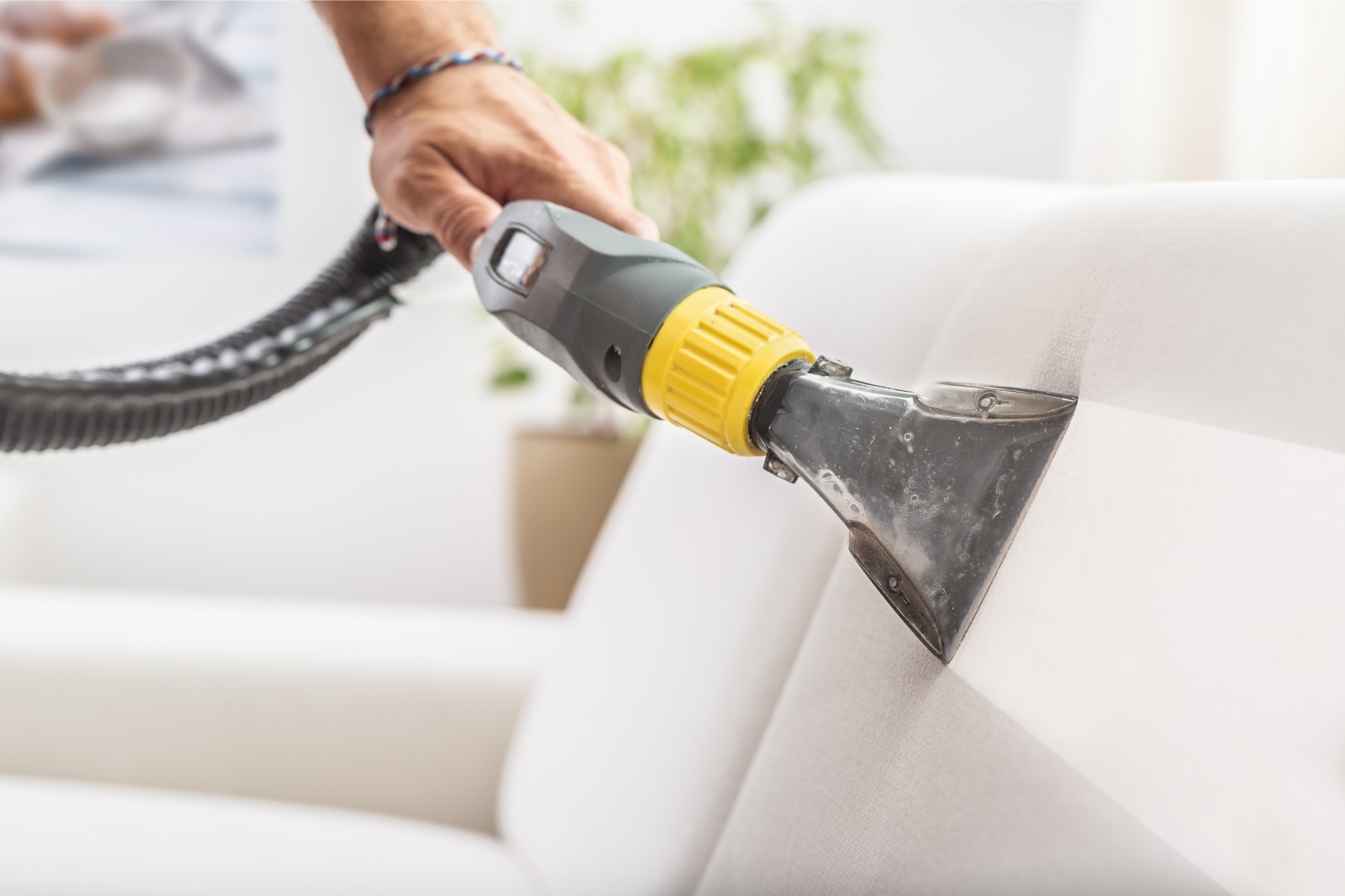 Person cleaning a white couch with a handheld upholstery cleaner in a well-lit room.