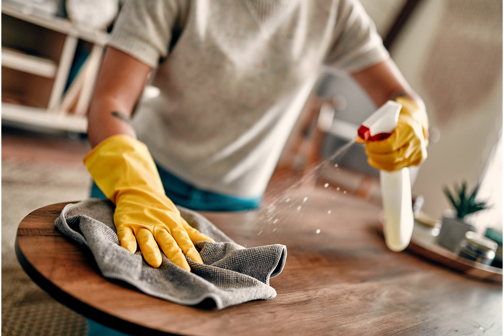 Person wearing yellow gloves spraying cleaner on a wooden table, wiping with a gray cloth.
