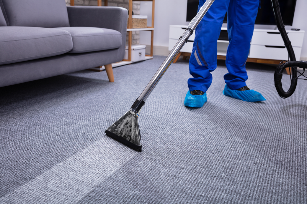 Person in blue cleaning a gray carpet with a vacuum, showing a clean strip.