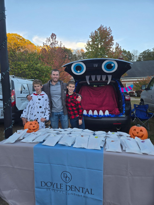 Three people posing beside a black monster-mouth display at a Doyl Dental Halloween booth with pumpkins.