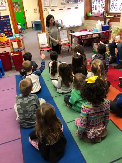Teacher reading to a group of seated children in a colorful classroom.