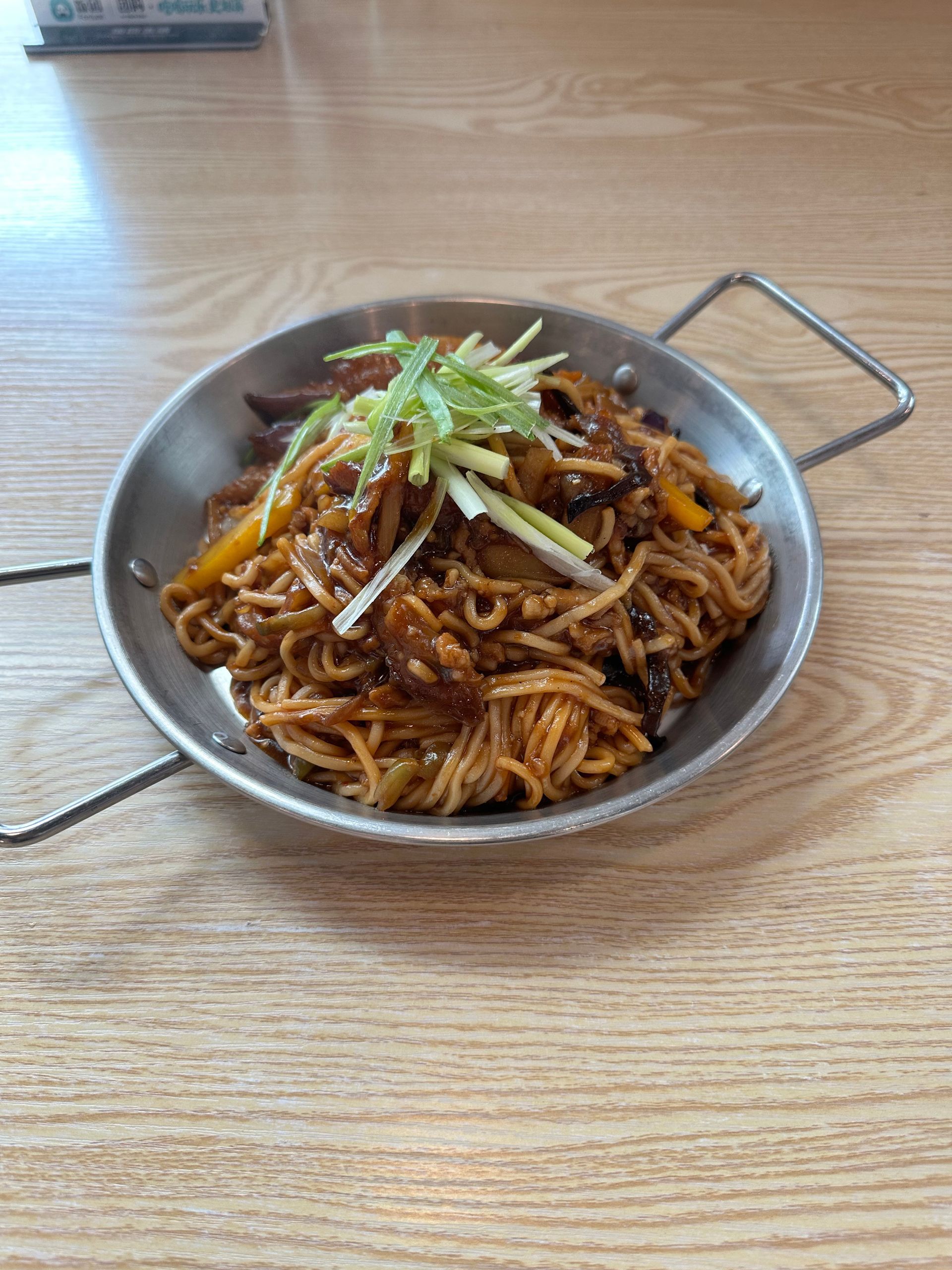 A metal bowl of spicy shredded stir-fry noodles topped with green onion on a wooden table.
