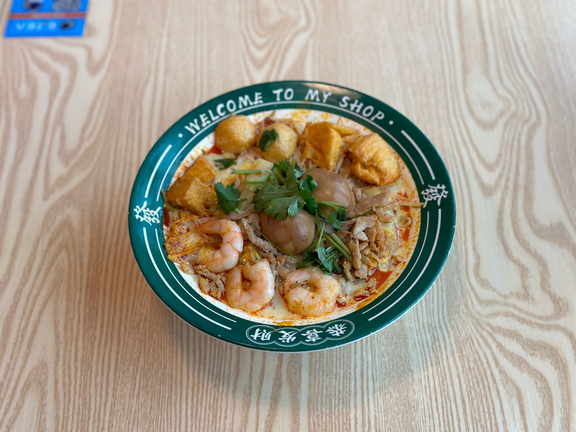 Bowl of noodle soup with shrimp and toppings on a green plate on a wood-grain table