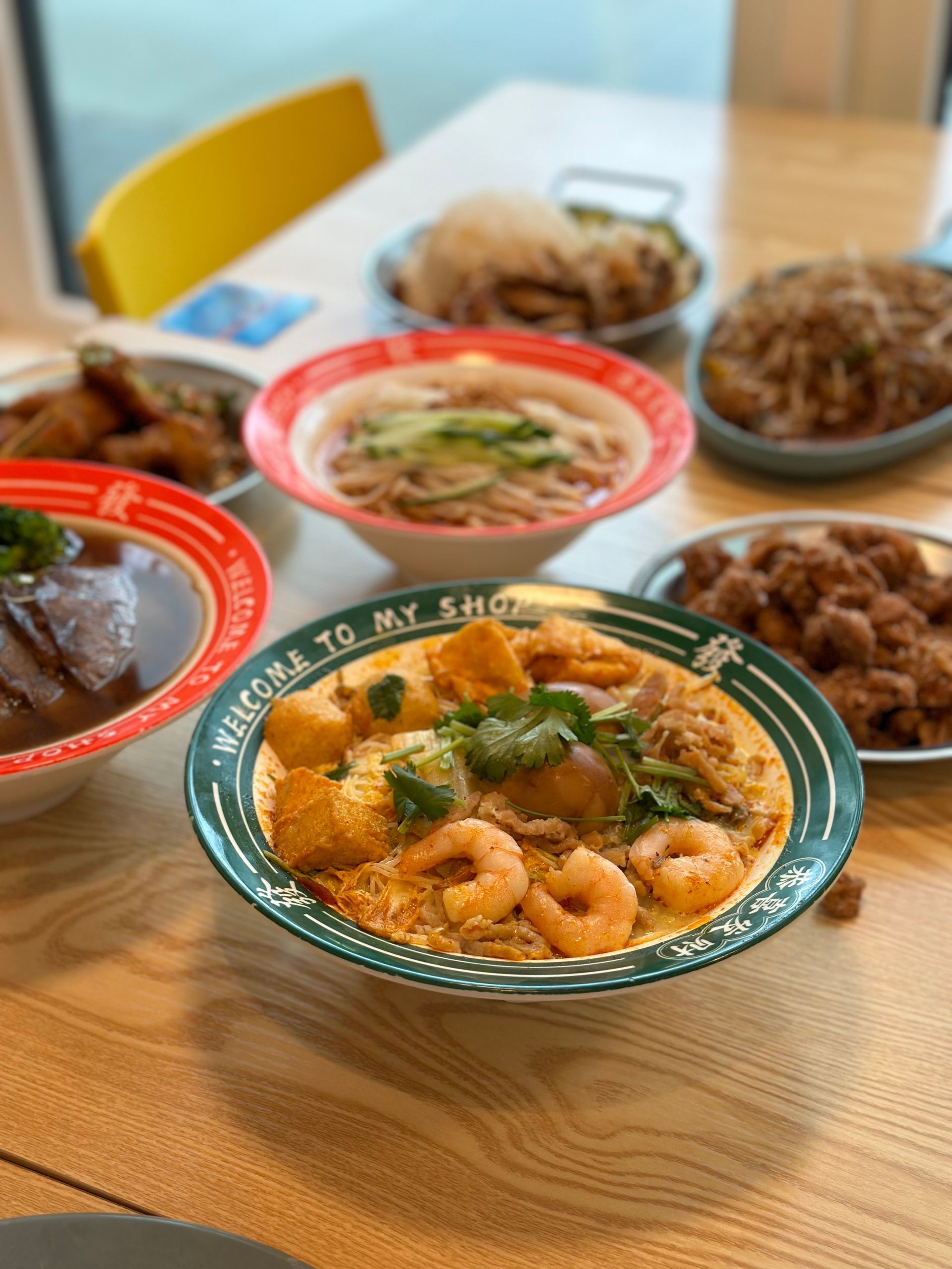 Several bowls of noodle soup and dishes on a wooden table in a bright dining room