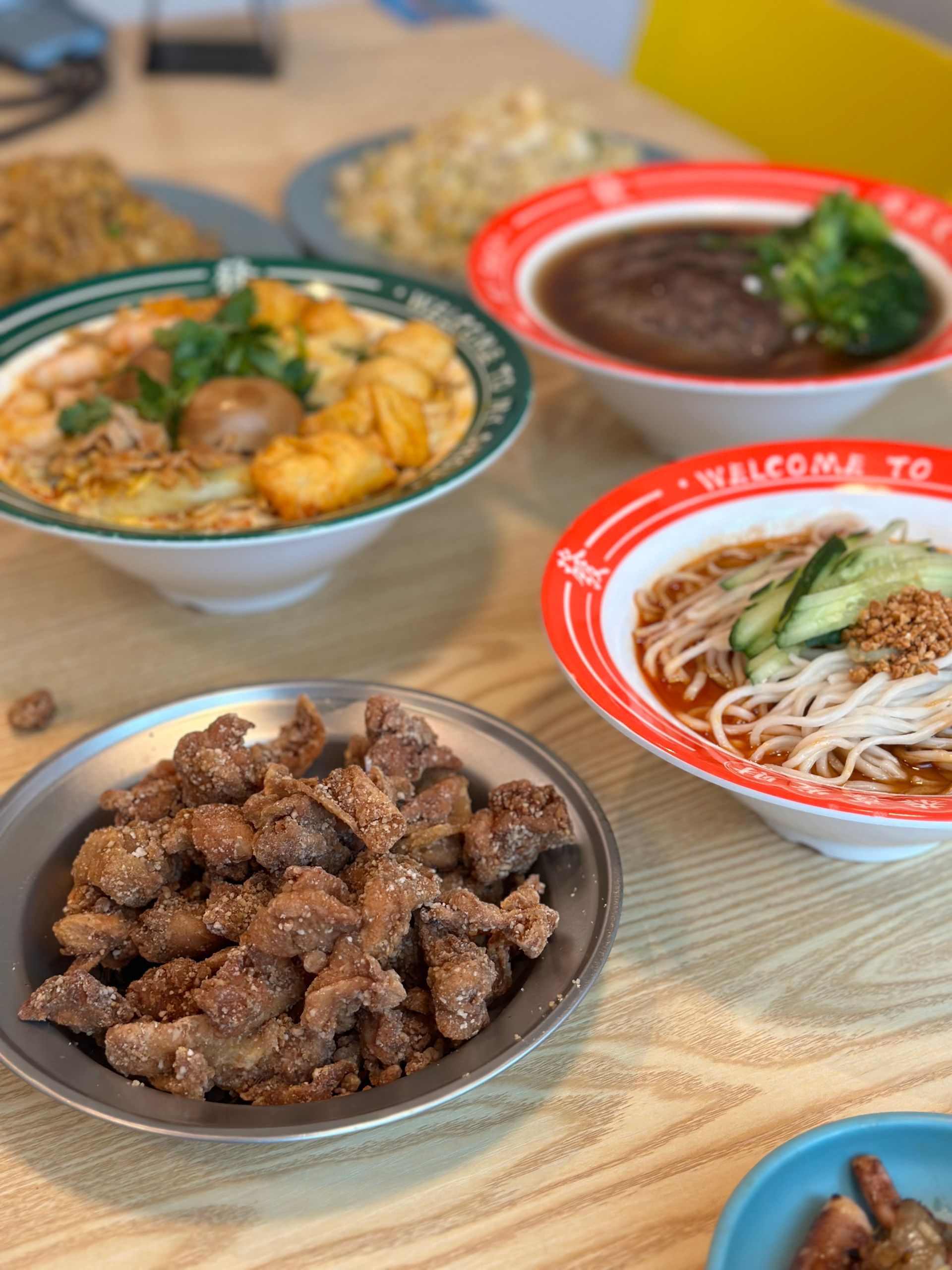 Four bowls of Asian dishes on a wooden table, including soup, meat, and noodles.