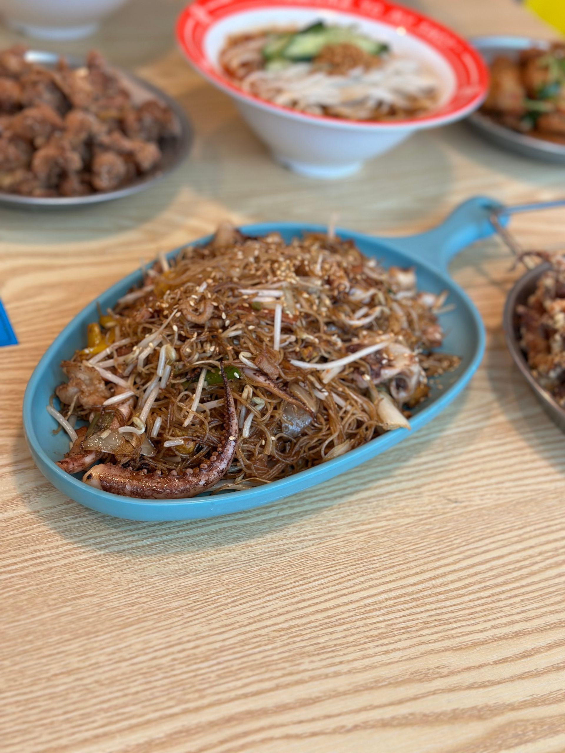 Blue plate of fried noodles topped with dried anchovies on a wooden table, with two dishes blurred in back