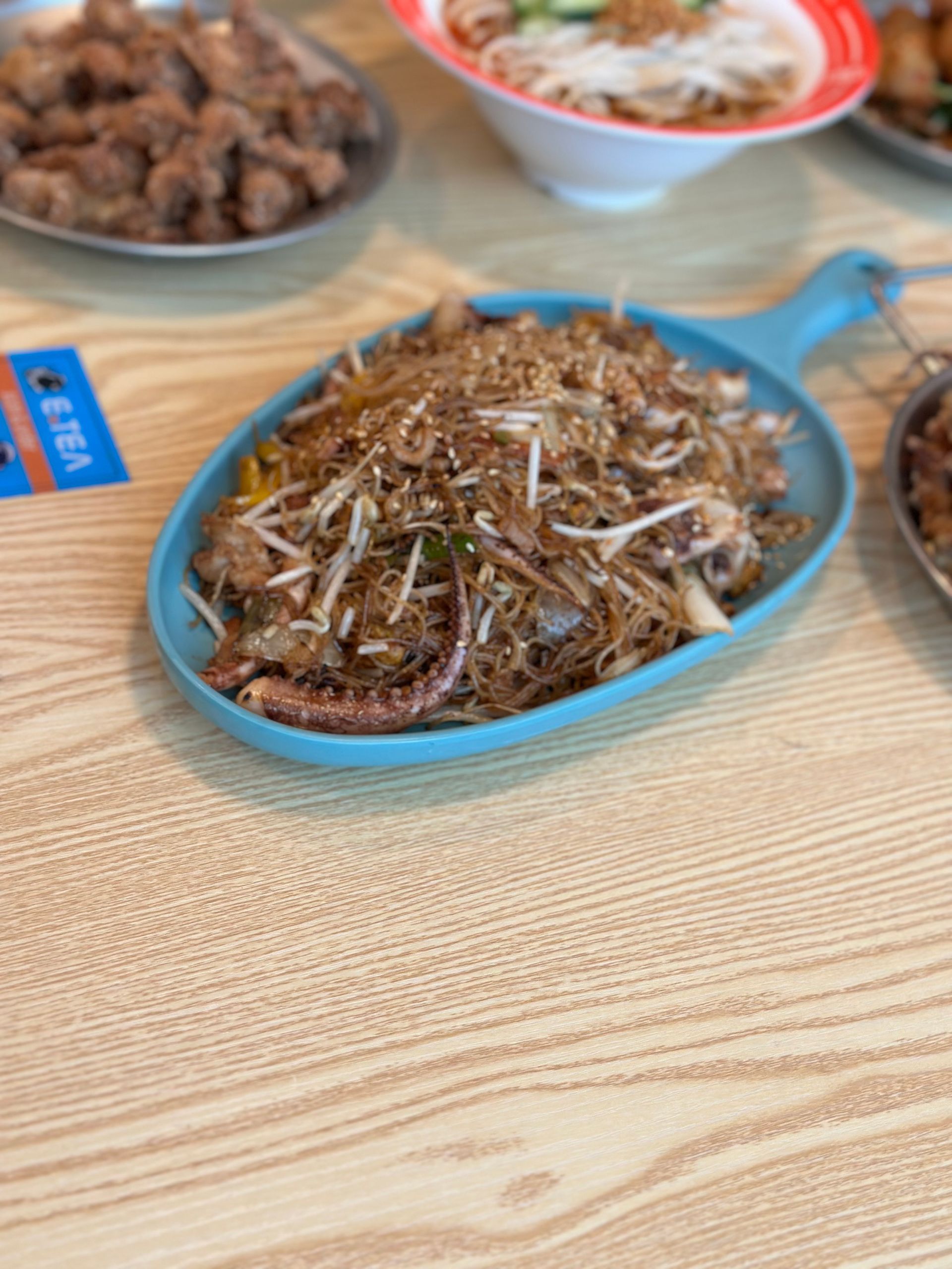 Blue dish of stir-fried noodles topped with sprouts on a wooden table, with other dishes blurred behind