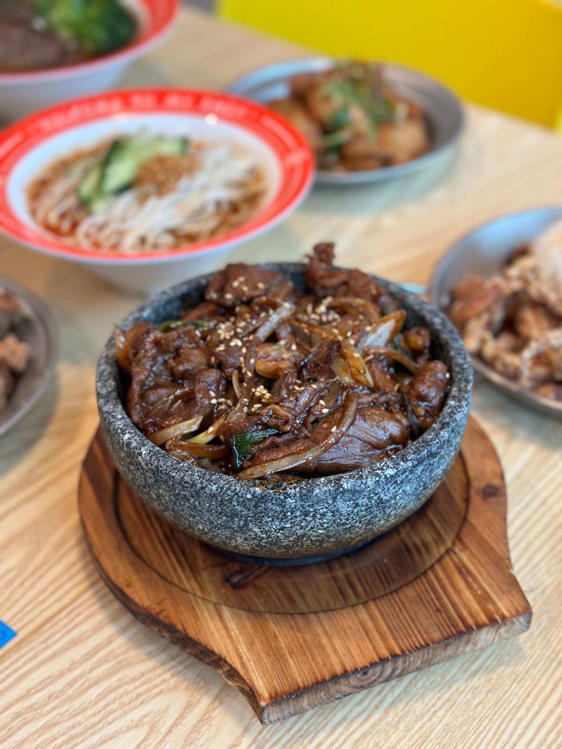 Stone bowl of braised meat and onions on a wooden board, with side dishes in the background.